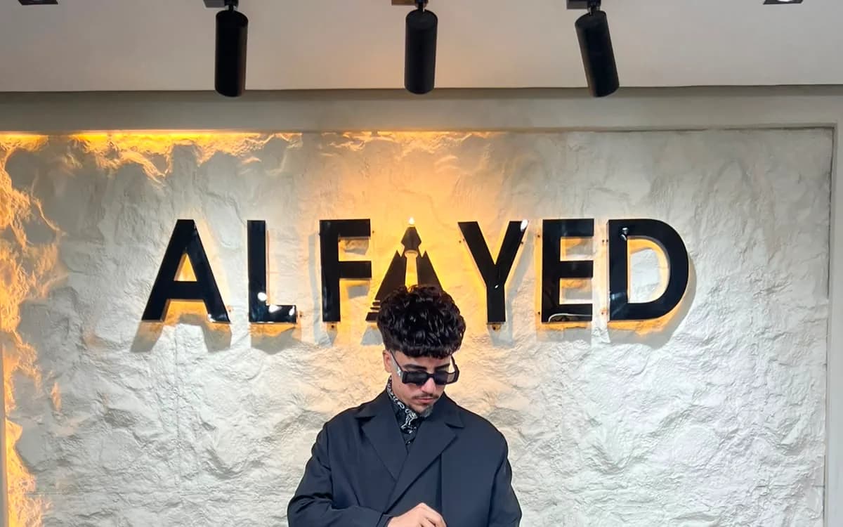A man in an oversized black suit, tailored wide, standing beneath the backlit ALFAYED sign in the Cairo house