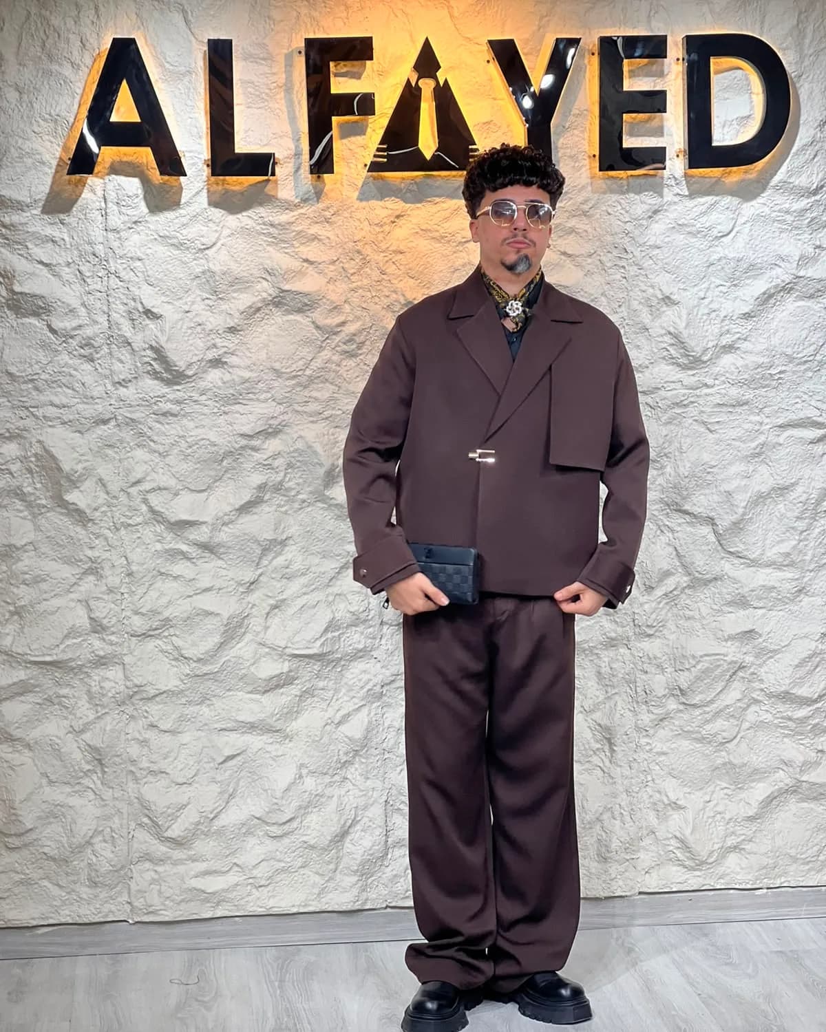 A man in an oversized dark-brown double-breasted suit, standing beneath the backlit ALFAYED sign in the Cairo house