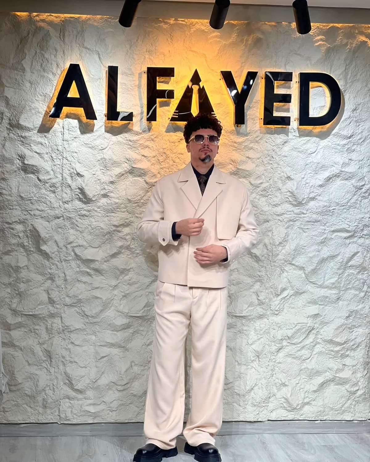 A man in a relaxed oversized cream suit, standing beneath the backlit ALFAYED sign in the Cairo house
