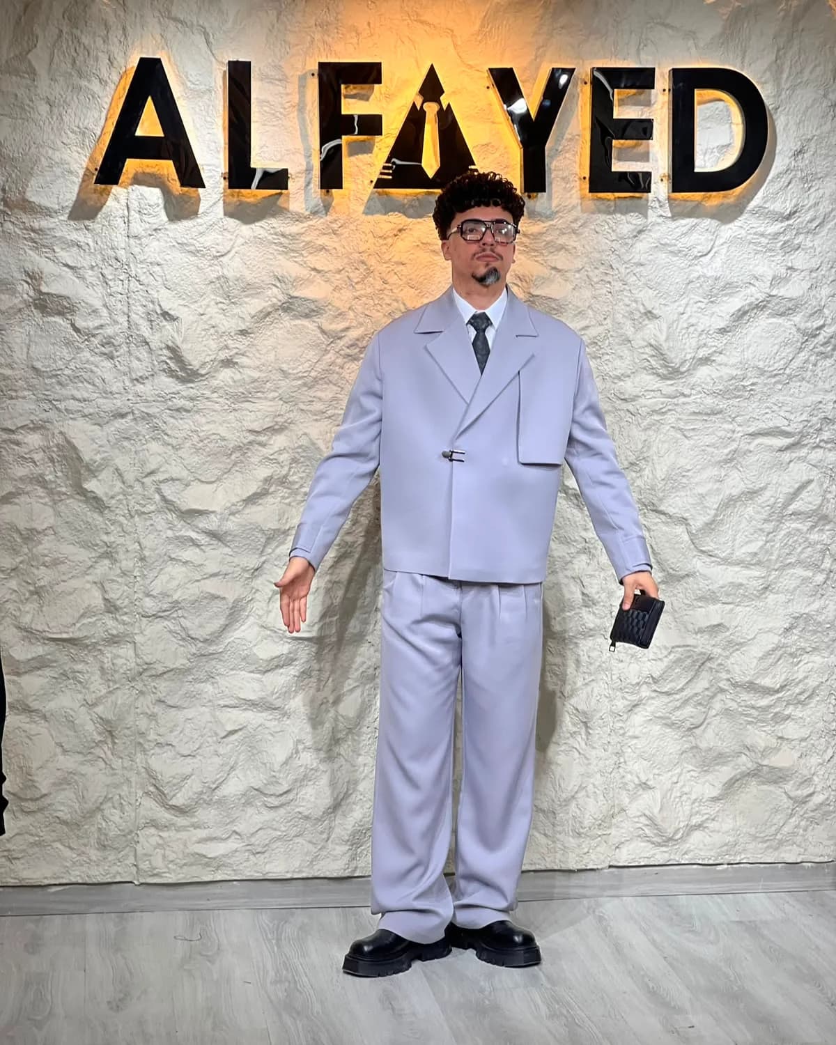 A man in an oversized pale-lilac suit with a soft shoulder, standing beneath the backlit ALFAYED sign in the Cairo house