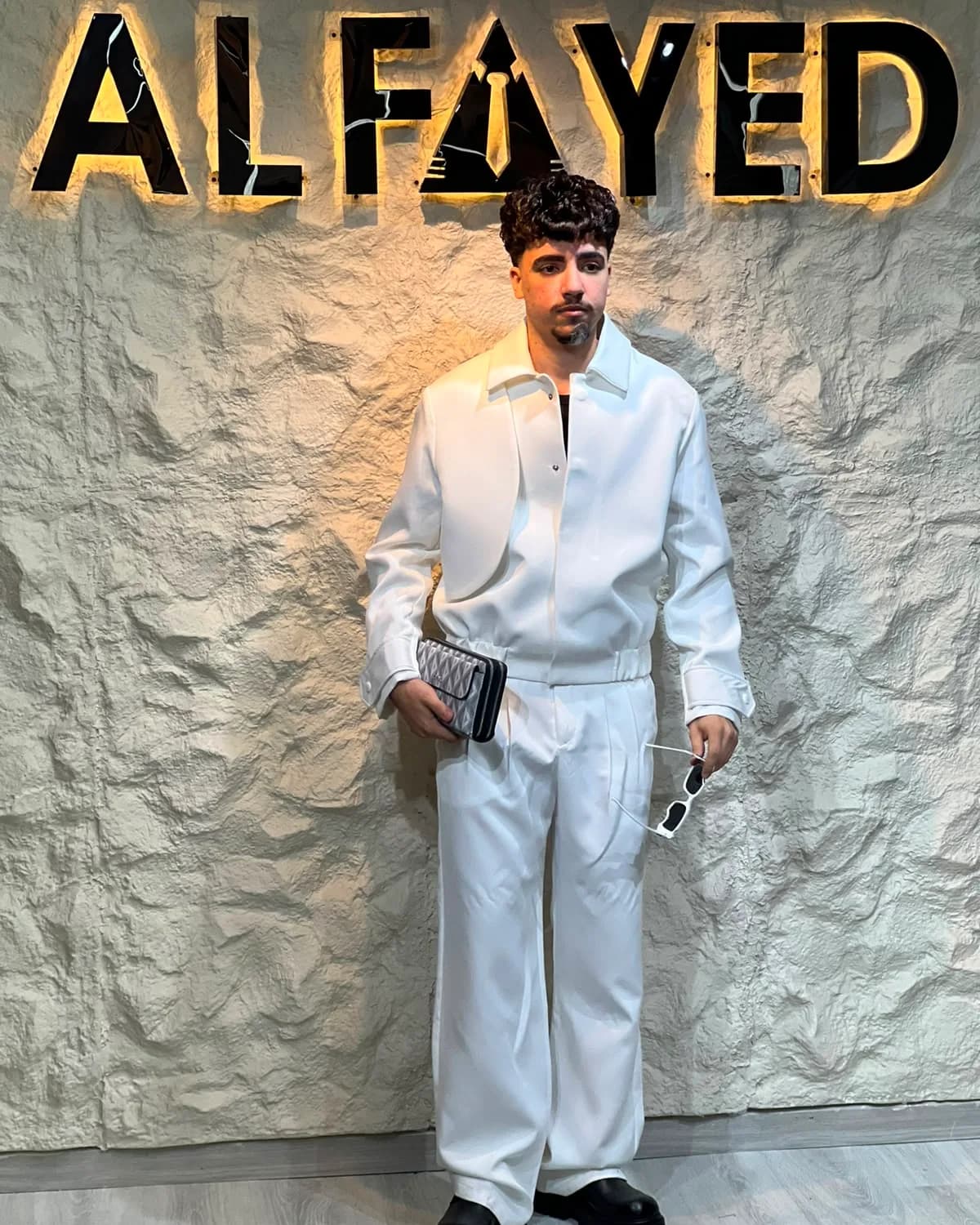 A man in an oversized white suit with an open collar, standing beneath the backlit ALFAYED sign in the Cairo house